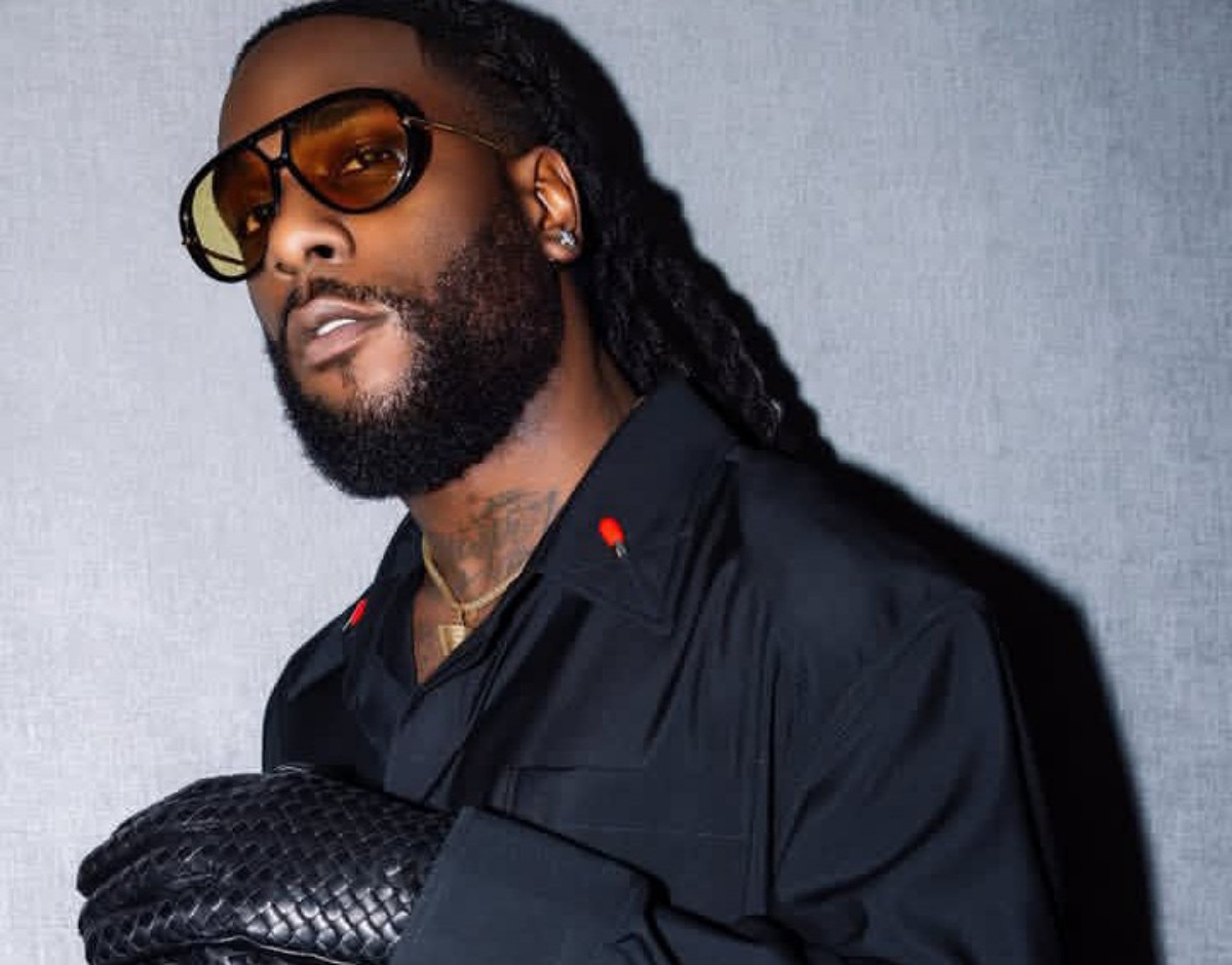 Man with a full beard and dreadlocks wearing amber-tinted sunglasses, a black shirt, a gold chain, and a textured black clutch resting on his arm.