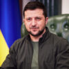Man with dark hair and beard sits in a green tufted chair, with a Ukrainian flag in the background.