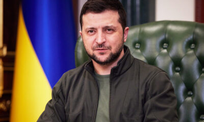 Man with dark hair and beard sits in a green tufted chair, with a Ukrainian flag in the background.