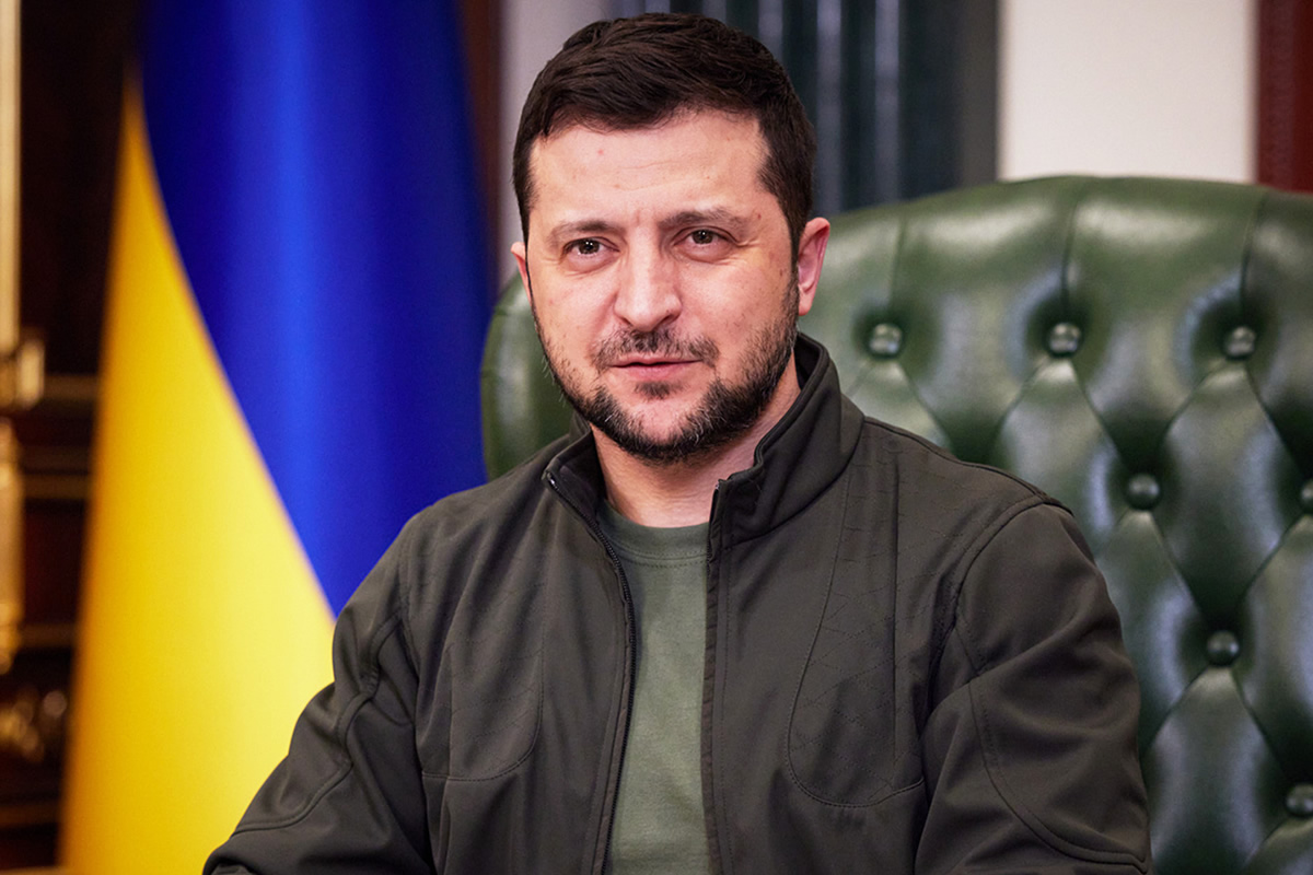 Man with dark hair and beard sits in a green tufted chair, with a Ukrainian flag in the background.