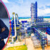 Smiling man in a dark blue shirt stands beside a large oil refinery complex by the seaside.