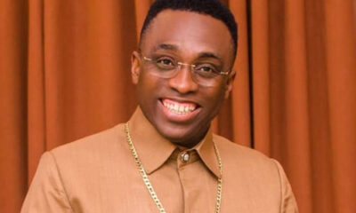 Smiling man with short hair wearing glasses, a tan shirt, and a gold chain posed against brown curtains.