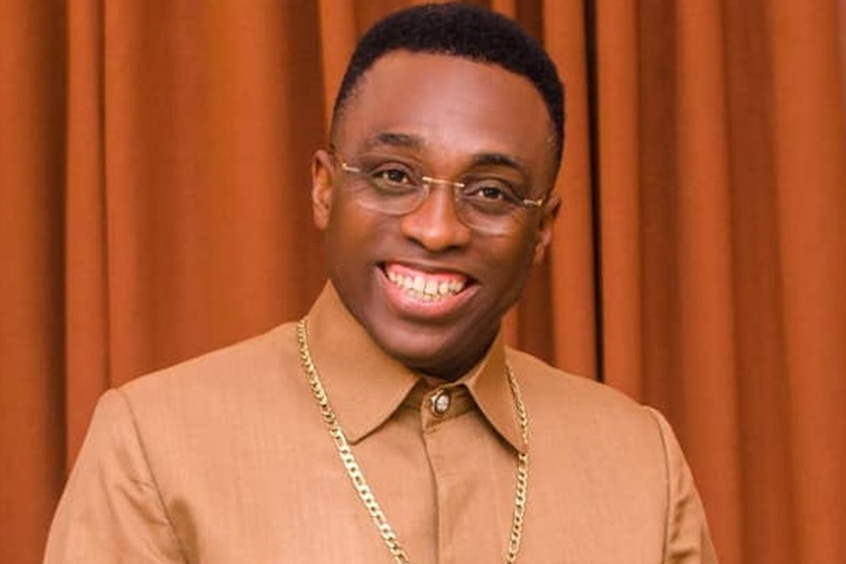 Smiling man with short hair wearing glasses, a tan shirt, and a gold chain posed against brown curtains.