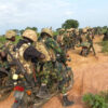 Armed soldiers in camouflage riding motorcycles in a line along a dusty rural road, rifles visible on their backs.