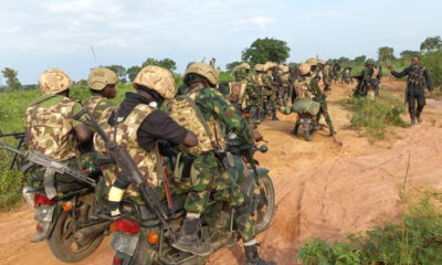 Armed soldiers in camouflage riding motorcycles in a line along a dusty rural road, rifles visible on their backs.