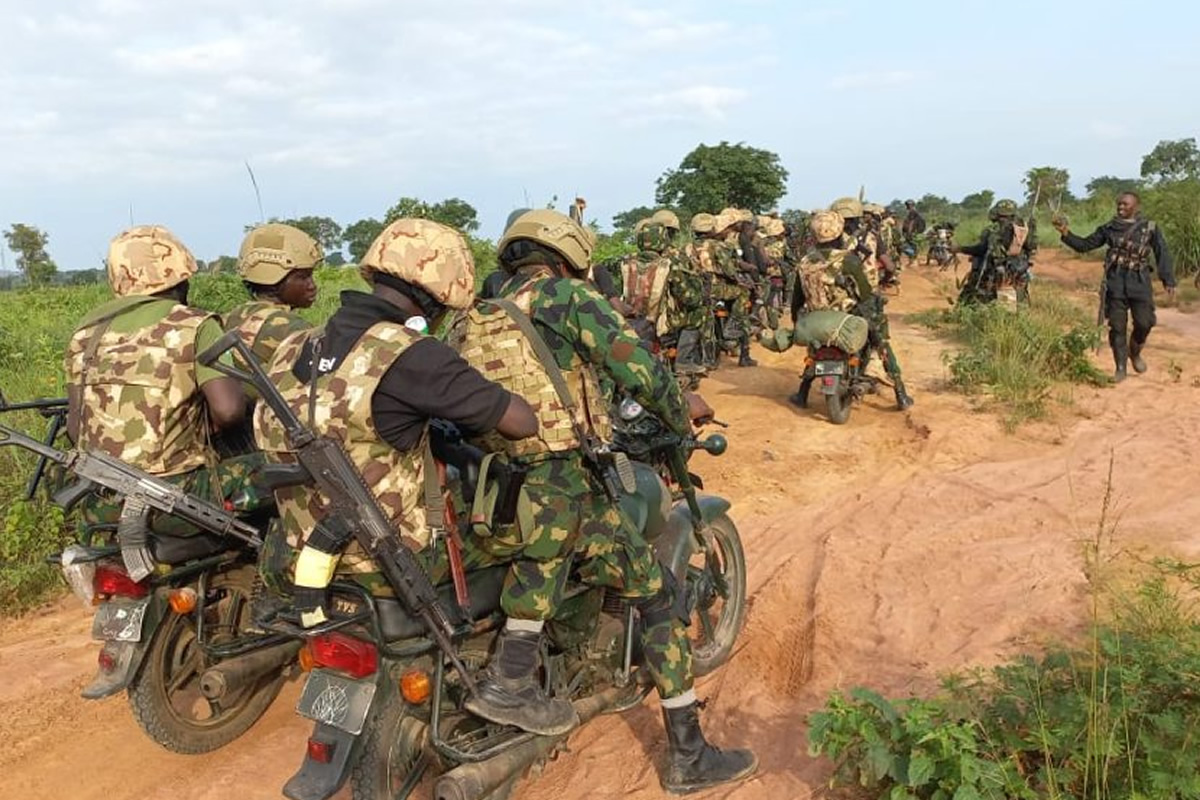 Armed soldiers in camouflage riding motorcycles in a line along a dusty rural road, rifles visible on their backs.