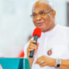 Man in white traditional attire and glasses speaks into a red microphone at a podium with a government seal on the front, smiling slightly.