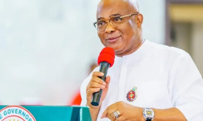 Man in white traditional attire and glasses speaks into a red microphone at a podium with a government seal on the front, smiling slightly.