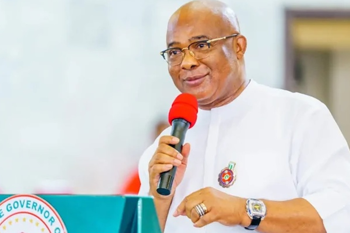 Man in white traditional attire and glasses speaks into a red microphone at a podium with a government seal on the front, smiling slightly.