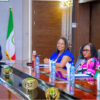 Four officials sit at a glossy conference table clapping; Nigerian flag and government seal visible in the background at a formal meeting.