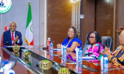 Four officials sit at a glossy conference table clapping; Nigerian flag and government seal visible in the background at a formal meeting.