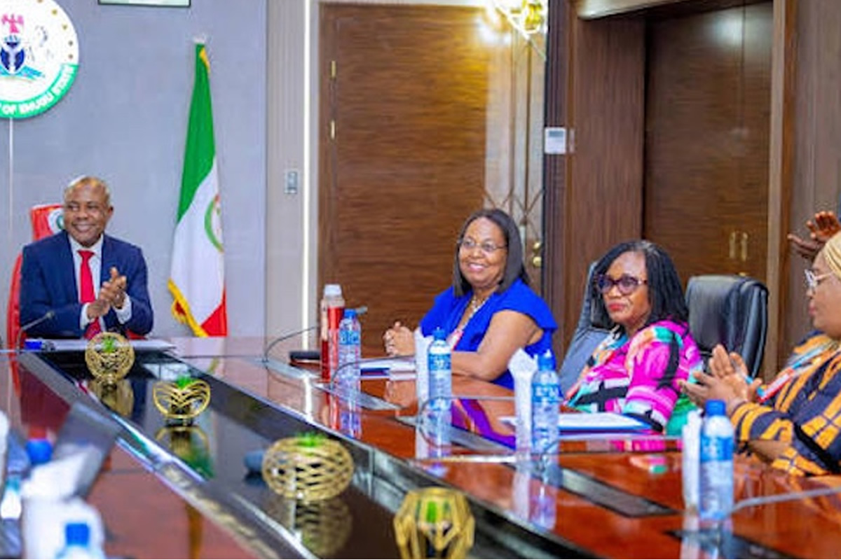 Four officials sit at a glossy conference table clapping; Nigerian flag and government seal visible in the background at a formal meeting.