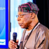 A Nigerian man in traditional attire speaks into a handheld microphone at a stage with a blue display and QR code beside him.