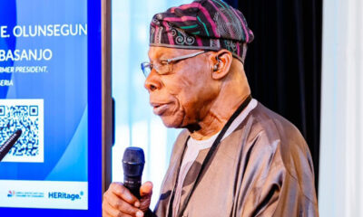 A Nigerian man in traditional attire speaks into a handheld microphone at a stage with a blue display and QR code beside him.