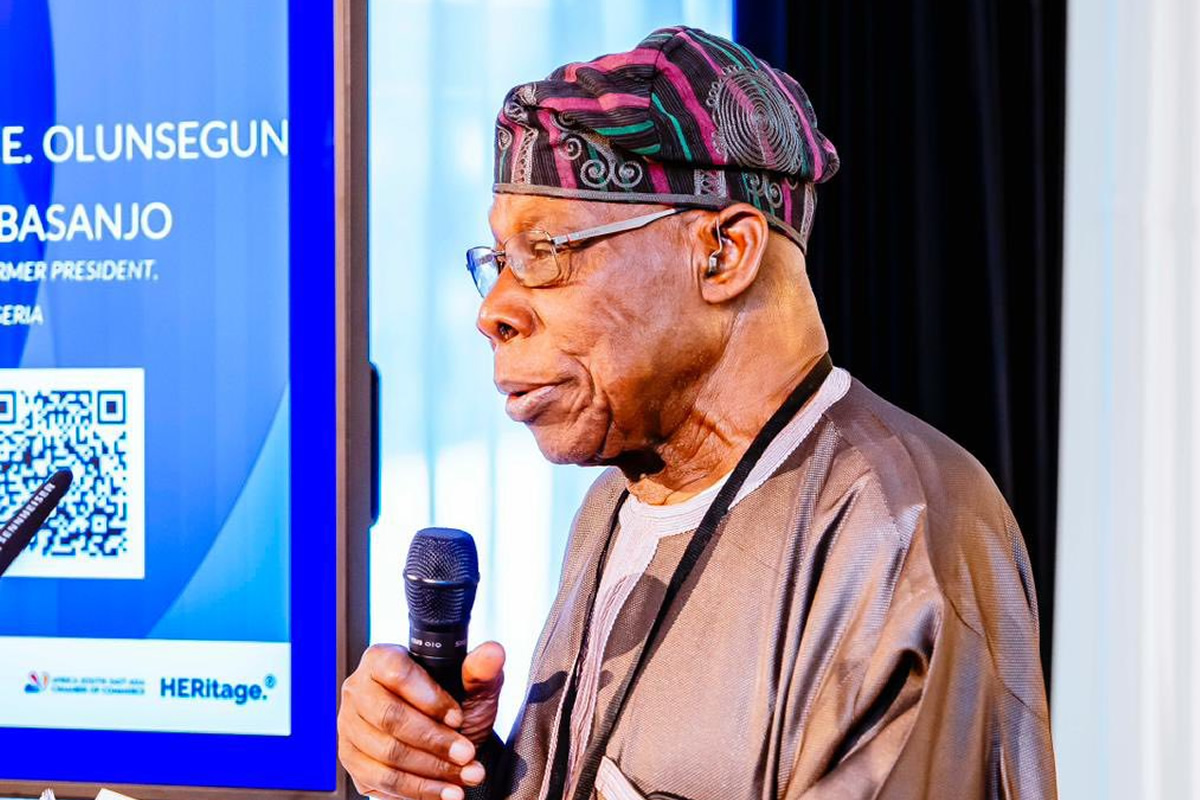 A Nigerian man in traditional attire speaks into a handheld microphone at a stage with a blue display and QR code beside him.