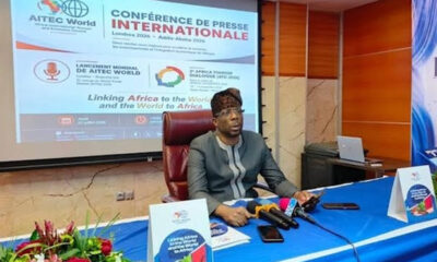 Man seated at a press conference desk with multiple microphones, a large projection screen behind showing 'CONFERENCE DE PRESSE INTERNATIONALE' and logos, and a flower arrangement in the foreground.