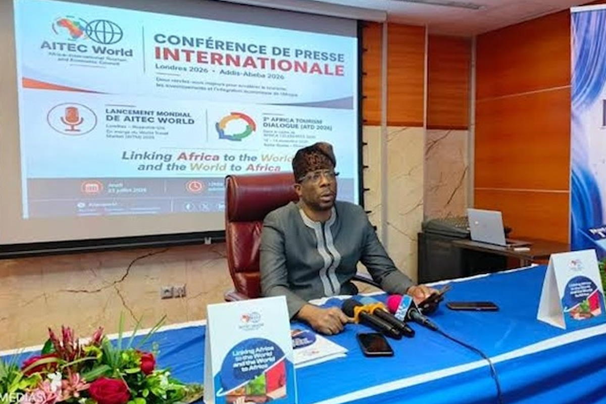 Man seated at a press conference desk with multiple microphones, a large projection screen behind showing 'CONFERENCE DE PRESSE INTERNATIONALE' and logos, and a flower arrangement in the foreground.