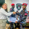 Woman in white blouse hands a certificate to a Nigerian police officer in camouflage during a formal ceremony; Nigeria Police insignia on the wall behind them.