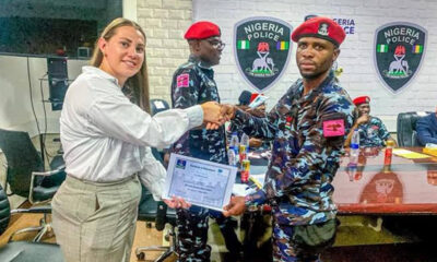 Woman in white blouse hands a certificate to a Nigerian police officer in camouflage during a formal ceremony; Nigeria Police insignia on the wall behind them.