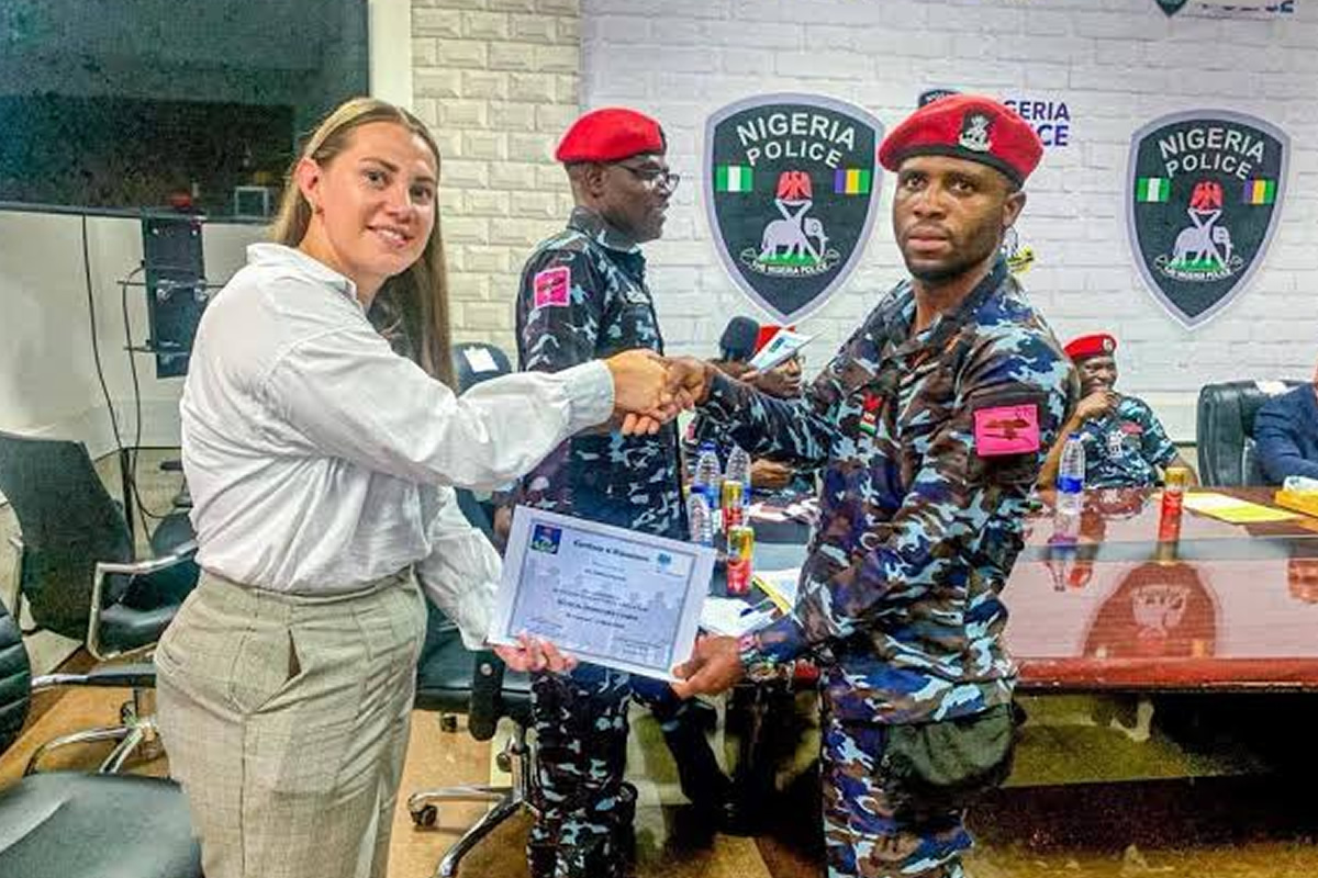 Woman in white blouse hands a certificate to a Nigerian police officer in camouflage during a formal ceremony; Nigeria Police insignia on the wall behind them.