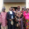Group of adults posing outside a building in formal attire; central man wears an ornate patterned suit and top hat.3Aefte 0:00:01