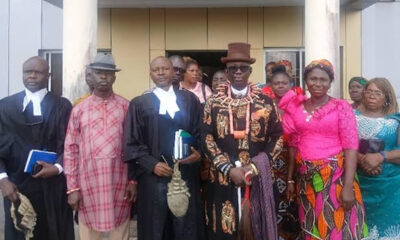 Group of adults posing outside a building in formal attire; central man wears an ornate patterned suit and top hat.3Aefte 0:00:01