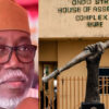 Portrait of a man wearing glasses and a brown cap, next to a bronze statue holding a torch in front of the Ondo State House of Assembly Complex, Akure, Nigeria.