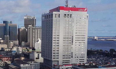 Tall grey skyscraper with a red UBA sign on top, part of a city skyline with water in the distance.