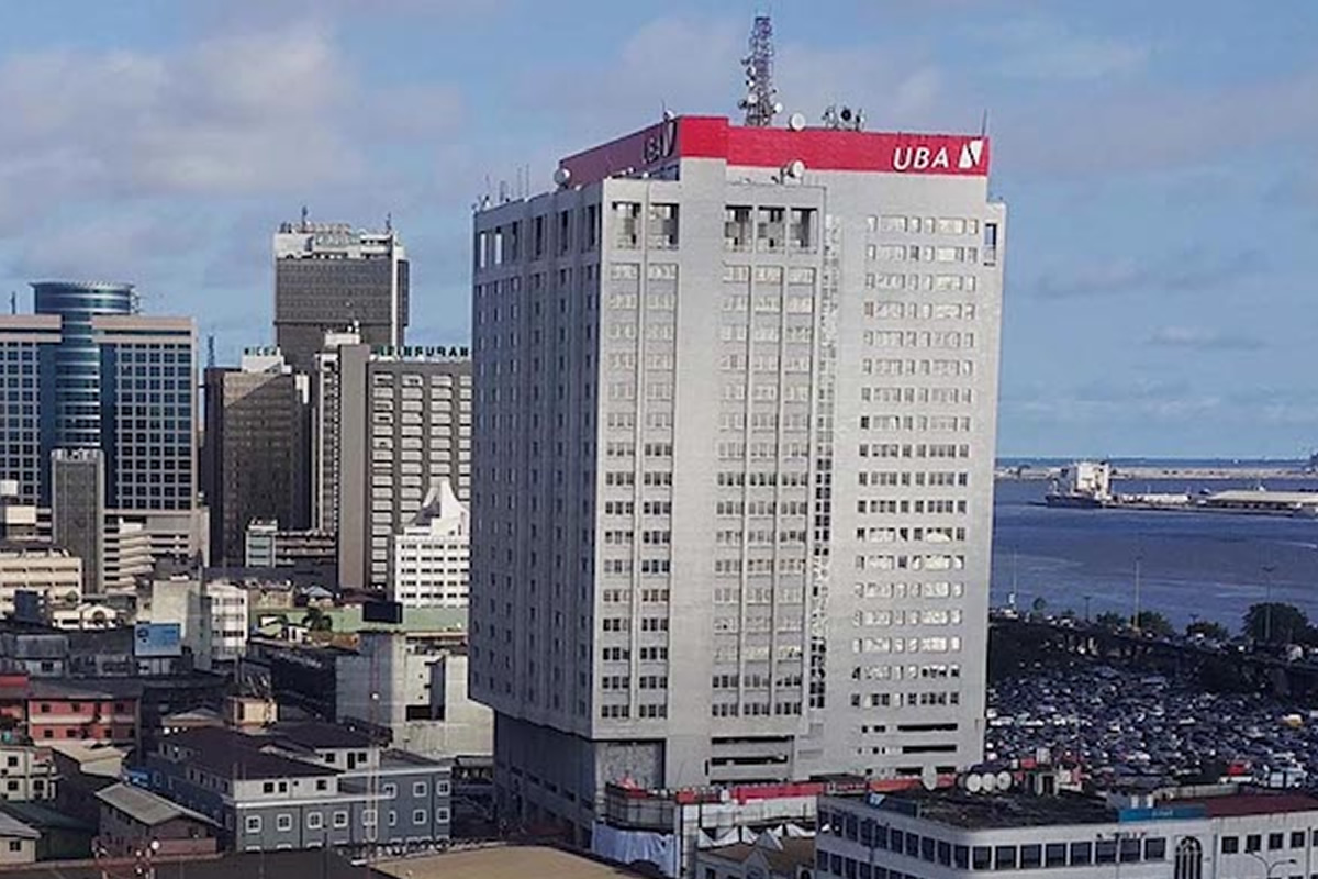 Tall grey skyscraper with a red UBA sign on top, part of a city skyline with water in the distance.