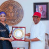 Two Nigerian officials exchange a framed award plaque in a government office, with the state seal on the wall behind them.