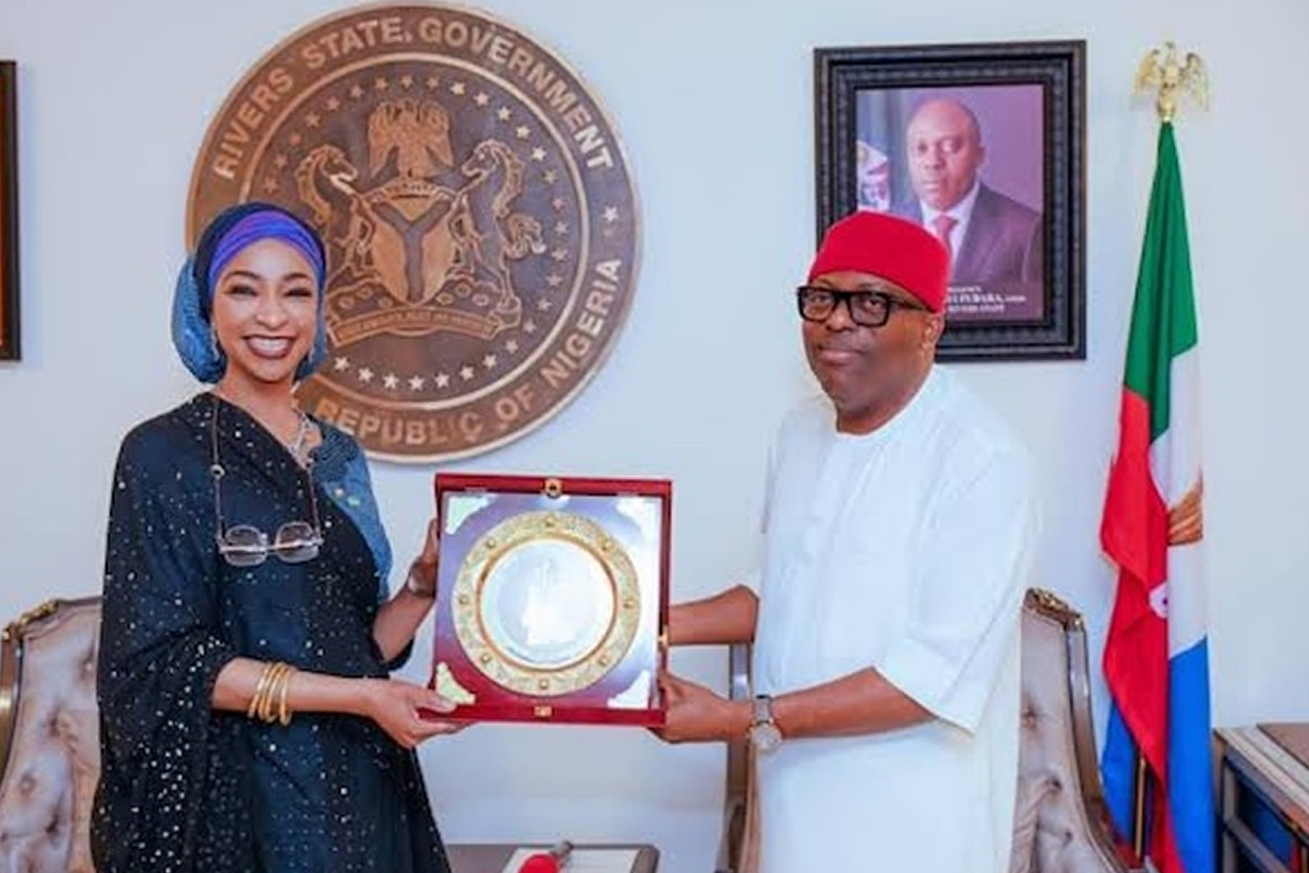 Two Nigerian officials exchange a framed award plaque in a government office, with the state seal on the wall behind them.