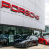 Porsche dealership exterior with red logo on a curved silver facade and several sports cars parked outside, including a black and a red model.