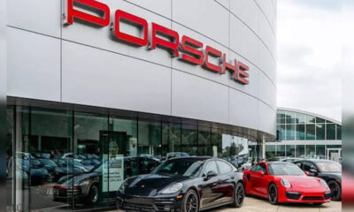 Porsche dealership exterior with red logo on a curved silver facade and several sports cars parked outside, including a black and a red model.