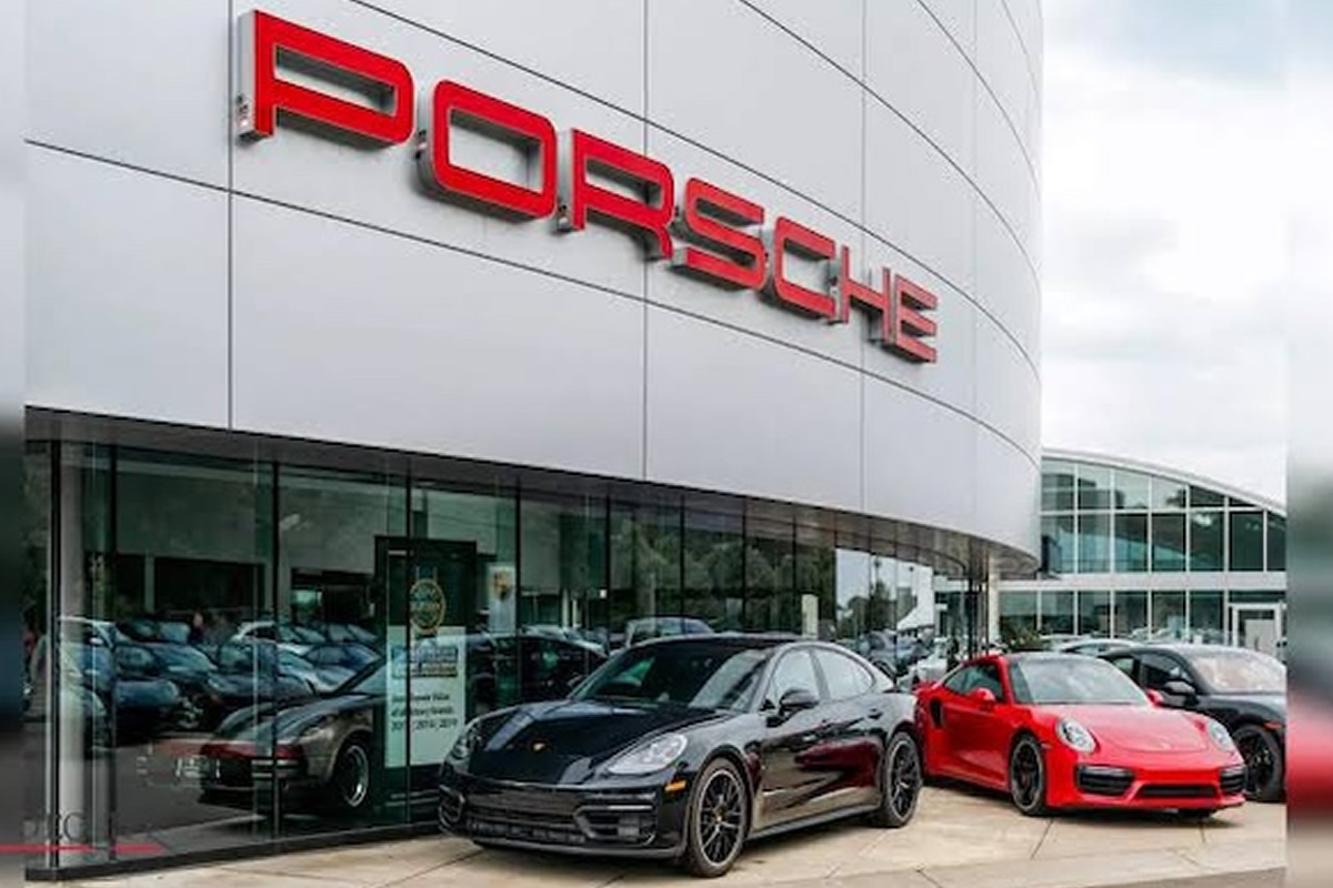 Porsche dealership exterior with red logo on a curved silver facade and several sports cars parked outside, including a black and a red model.