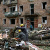 Rescue workers in yellow helmets clearing debris from a collapsed street in front of a damaged brick apartment building.