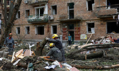 Rescue workers in yellow helmets clearing debris from a collapsed street in front of a damaged brick apartment building.