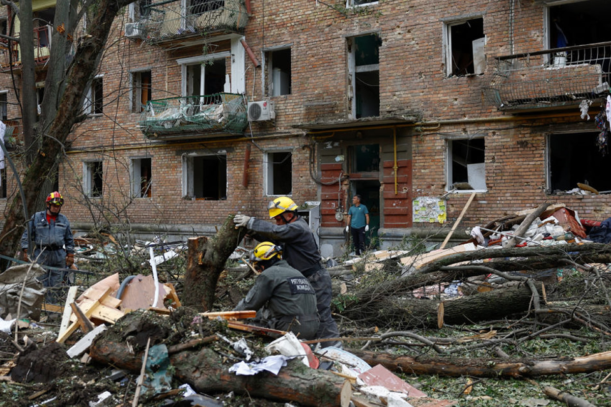 Rescue workers in yellow helmets clearing debris from a collapsed street in front of a damaged brick apartment building.
