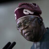 Older man wearing a maroon headwrap and round glasses speaks into two microphones.