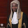 Male judge wearing a traditional white wig and black robes with gold trim in a courtroom setting.
