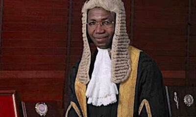 Male judge wearing a traditional white wig and black robes with gold trim in a courtroom setting.
