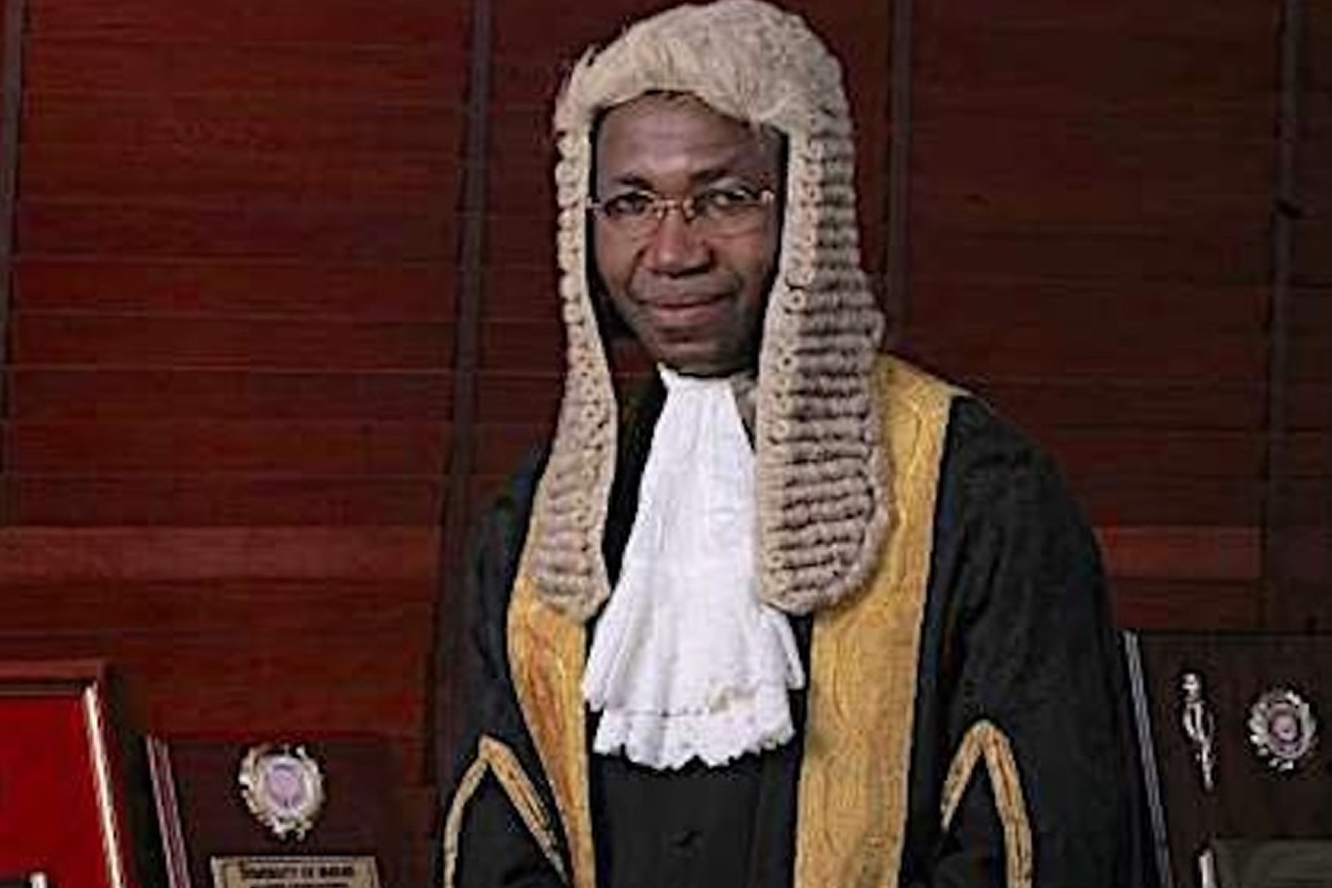 Male judge wearing a traditional white wig and black robes with gold trim in a courtroom setting.