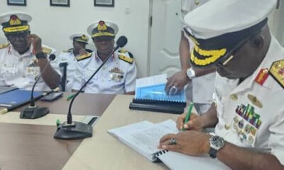 Naval officers in white dress uniforms signing documents at a conference table with microphones nearby.