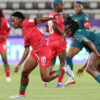 Soccer players in red and teal jerseys chase the ball on a green field during a match, one red player sprinting ahead.
