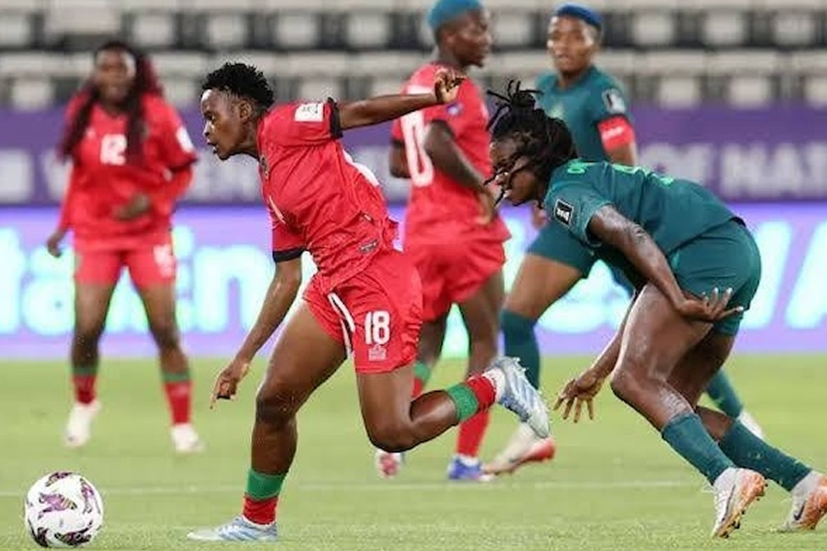 Soccer players in red and teal jerseys chase the ball on a green field during a match, one red player sprinting ahead.