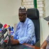 Three men sit at a table with numerous microphones in front of them, speaking at a press conference in an office setting.