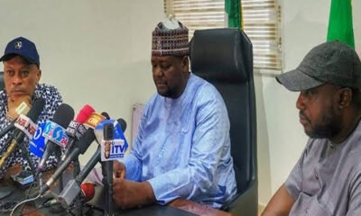 Three men sit at a table with numerous microphones in front of them, speaking at a press conference in an office setting.