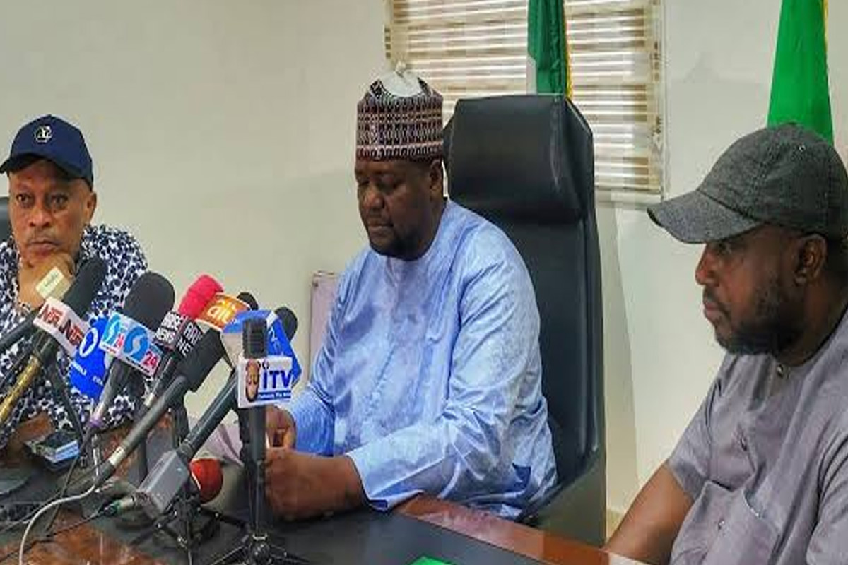 Three men sit at a table with numerous microphones in front of them, speaking at a press conference in an office setting.