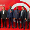 Six men in dark suits stand side by side on a red carpet at a corporate event, posing in front of a red backdrop with a white circular logo.