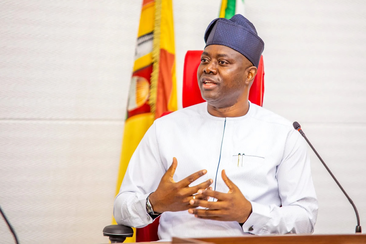 Man in light blue traditional attire and cap speaks into a microphone during a formal meeting.