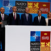 Group of officials in suits standing behind a podium at a NATO summit in Madrid 2022. Signs read NATO/OTAN and Madrid summit.